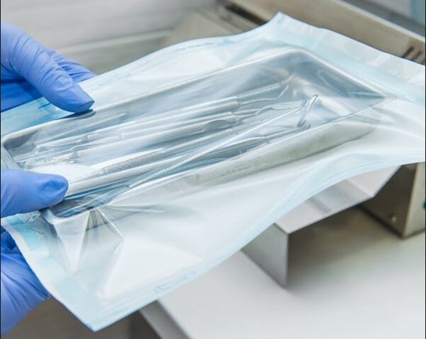 Gloved hands holding a sealed sterile pouch containing stainless steel medical instruments